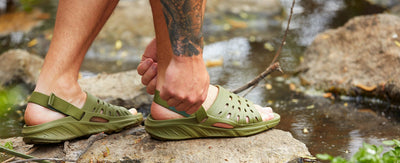 man adjusting the rear strap on his joybees trekking slides while standing on a rock in a creek